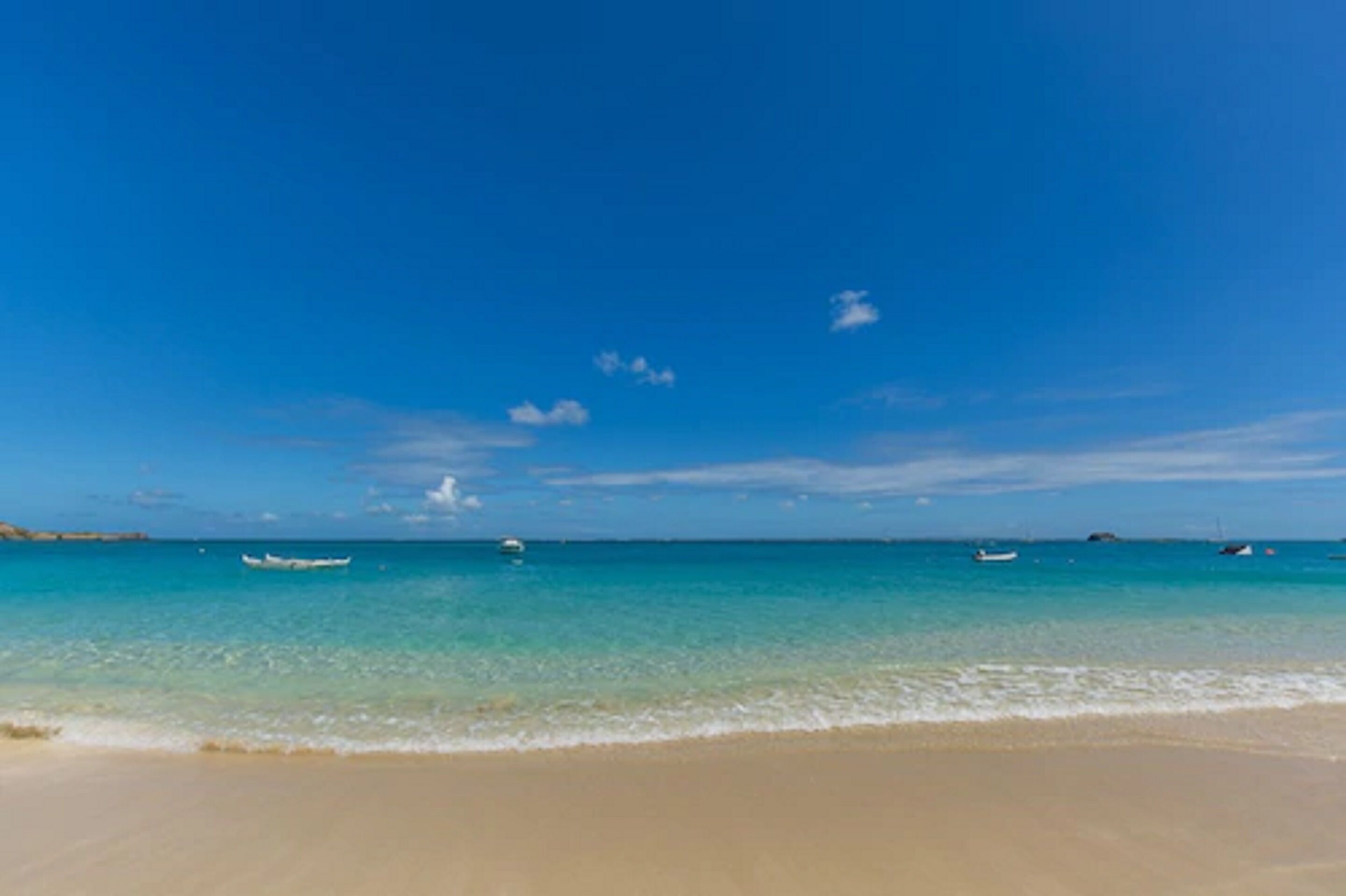 Una playa con agua turquesa cristalina, una orilla arenosa y algunos barcos a lo lejos.