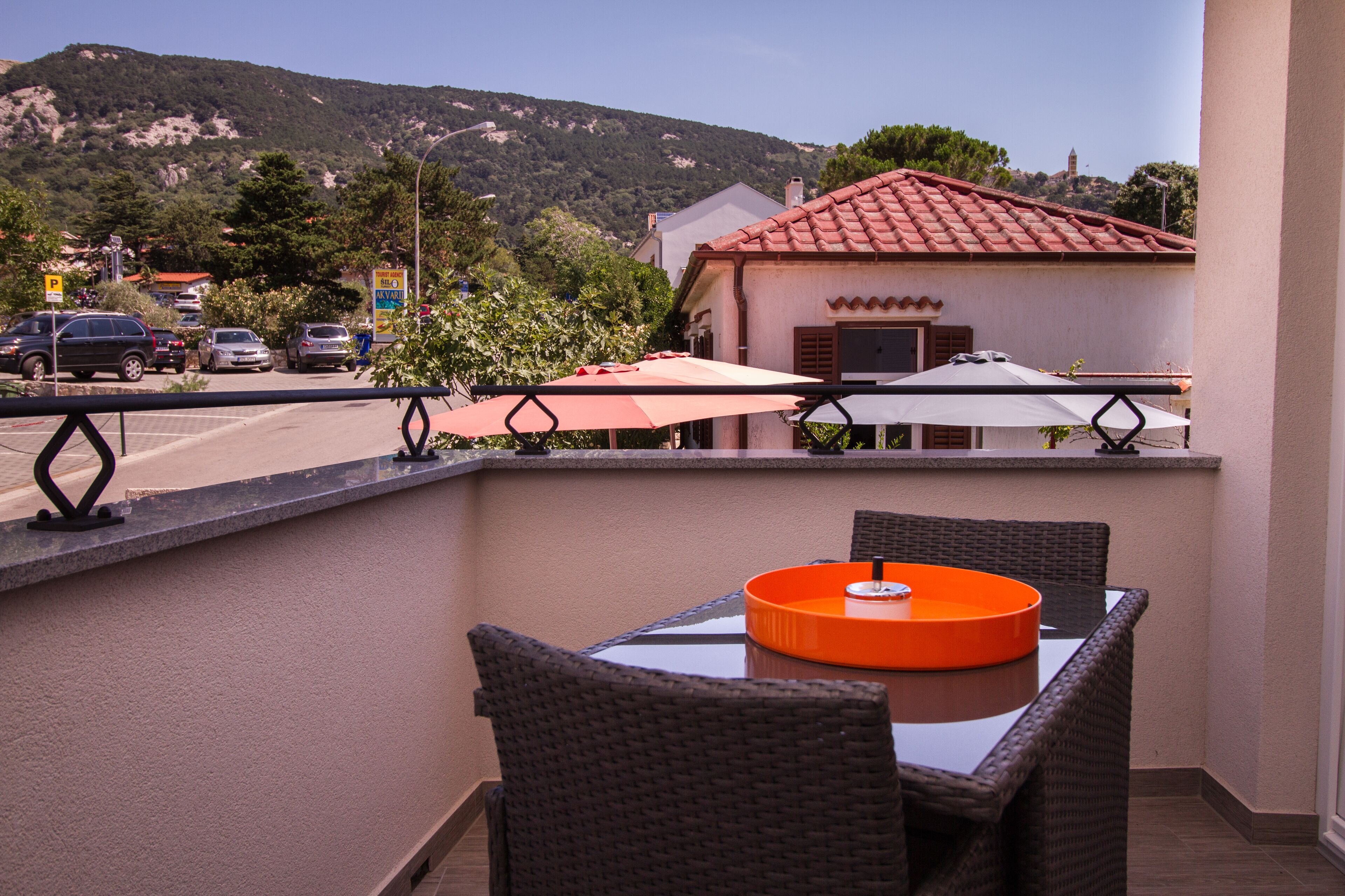 Un balcon avec des fauteuils en osier et une table, donnant sur une rue où sont garées des voitures et sur un immeuble avec un toit rouge.