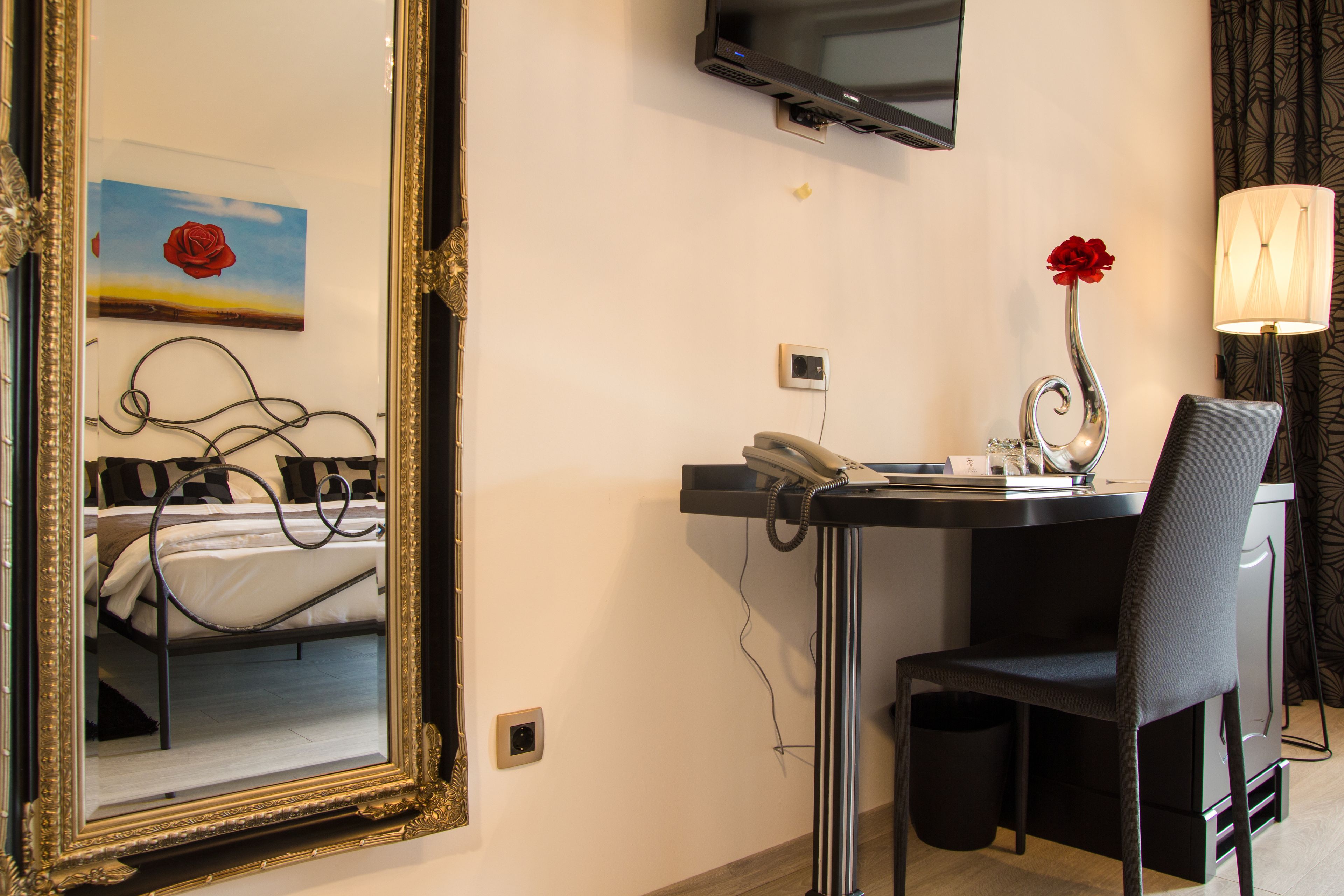 A hotel room with a bed, a desk with a chair, a TV mounted on the wall, and a vase with a red flower on the desk.