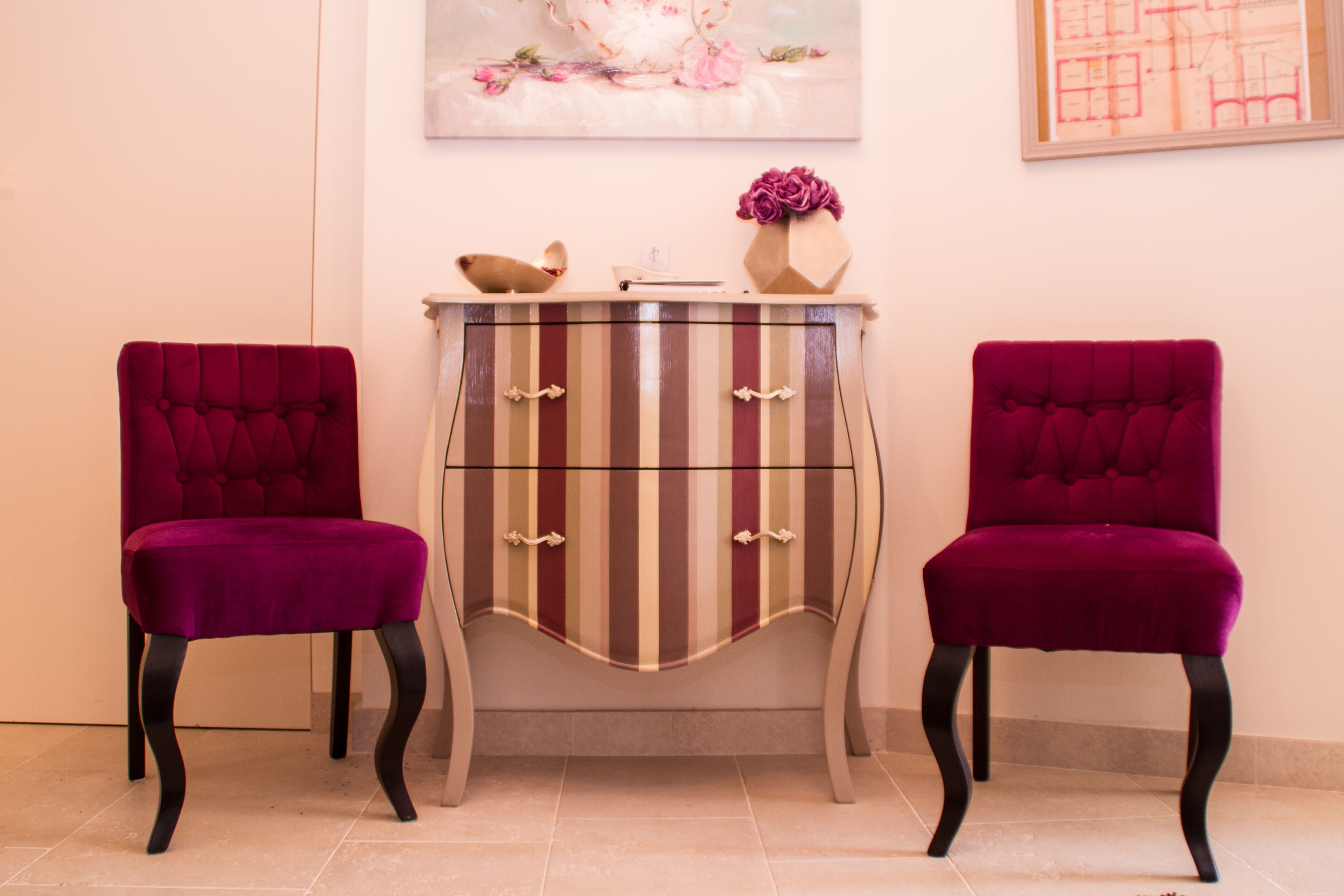 Deux fauteuils moelleux de couleur violette, un meuble à rayures et un vase avec des fleurs roses, le tout placé contre un mur.