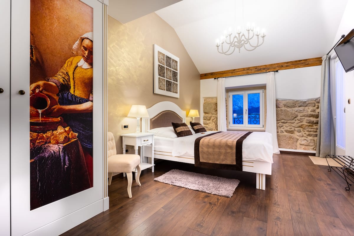 A bedroom with a bed, bedside tables, a nightstand, a window with a view, and a painting on the wall.