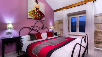 Une chambre à coucher avec un lit, deux tables de chevet, une commode, un tableau, une fenêtre avec des rideaux et un mur en pierre.