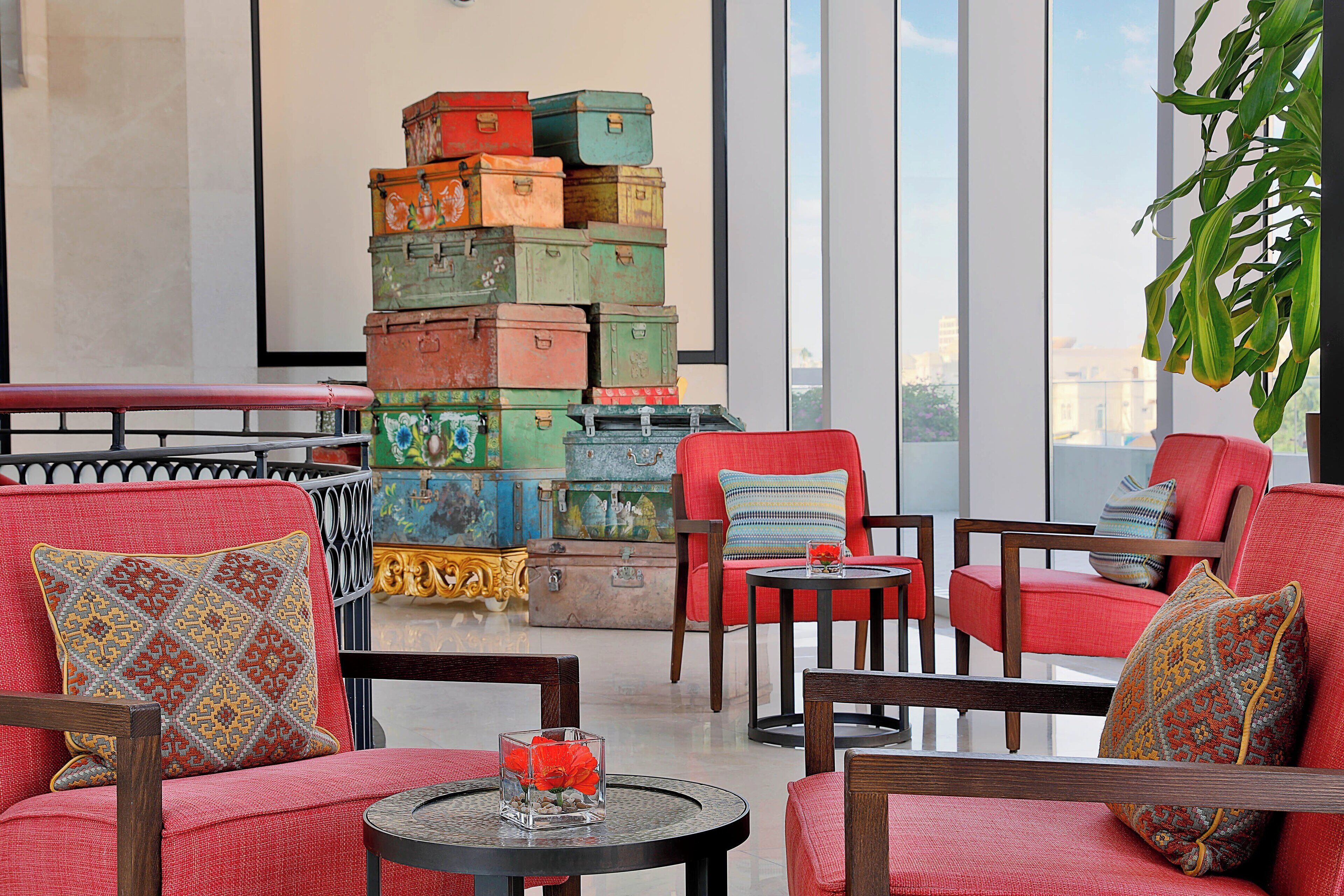 Un salón moderno con sillones tapizados en rojo, una pila de maletas antiguas y vistas de la ciudad a través de amplias ventanas.