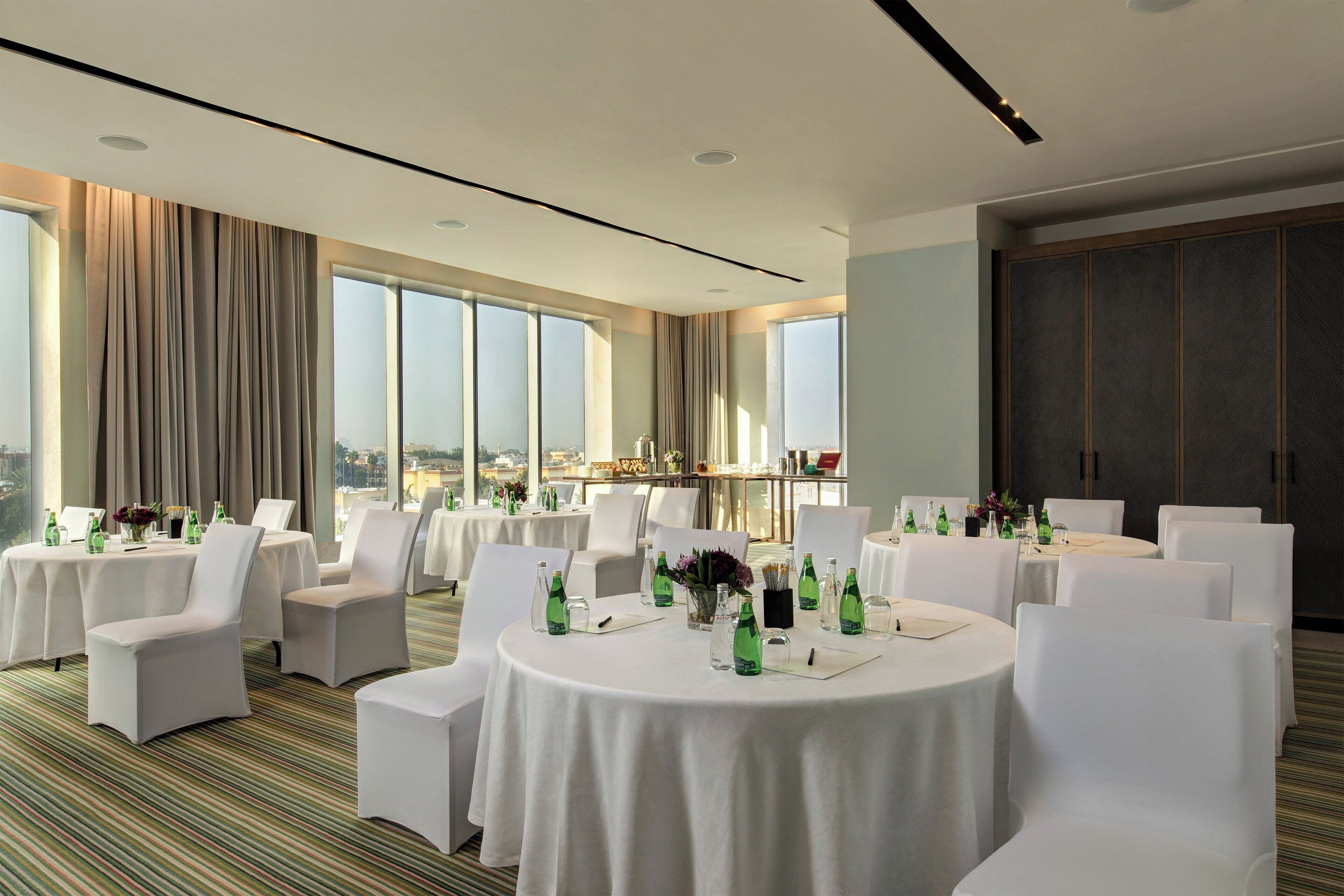 A hotel conference room with round tables, white chairs, and large windows offering a city view.