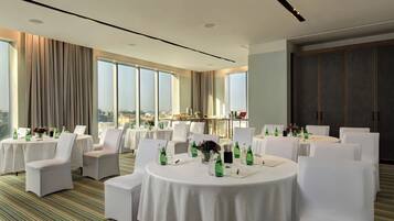 A hotel conference room with round tables, white chairs, and large windows offering a city view.
