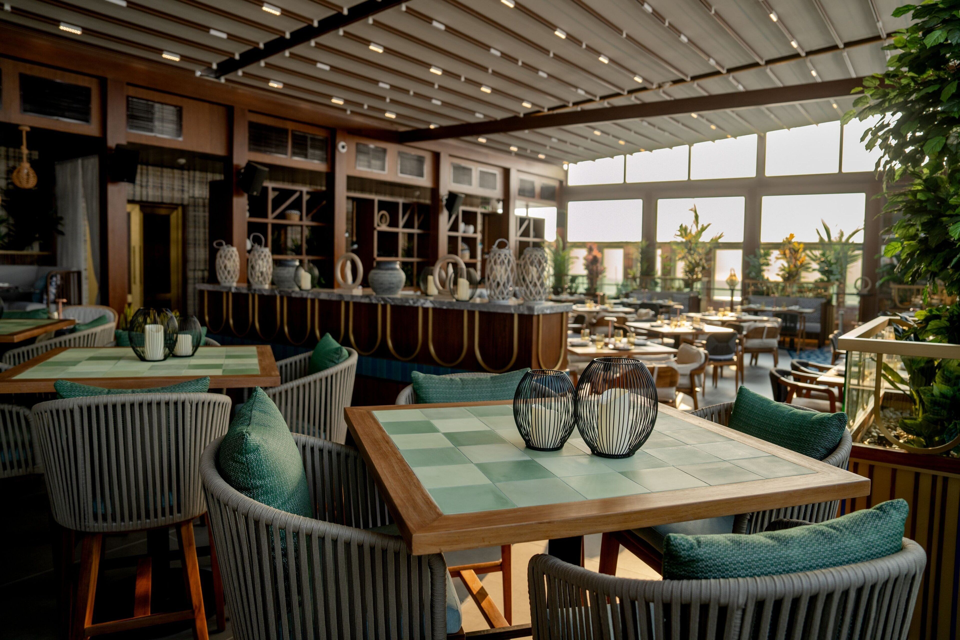 Ein Restaurant mit Korbstühlen, karierten Tischdecken und Blick ins Freie durch große Fenster.