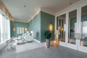 A modern hotel lobby with a teal wall, a reception desk, a seating area with white chairs, a large potted plant, and a glass door entrance.
