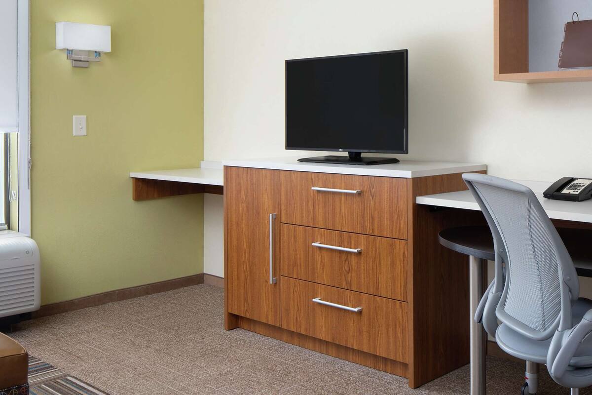 Une chambre d’hôtel équipée d’un bureau, d’une chaise, d’une télévision, d’un téléphone et d’une petite étagère sur laquelle se trouve un sac d’accueil.