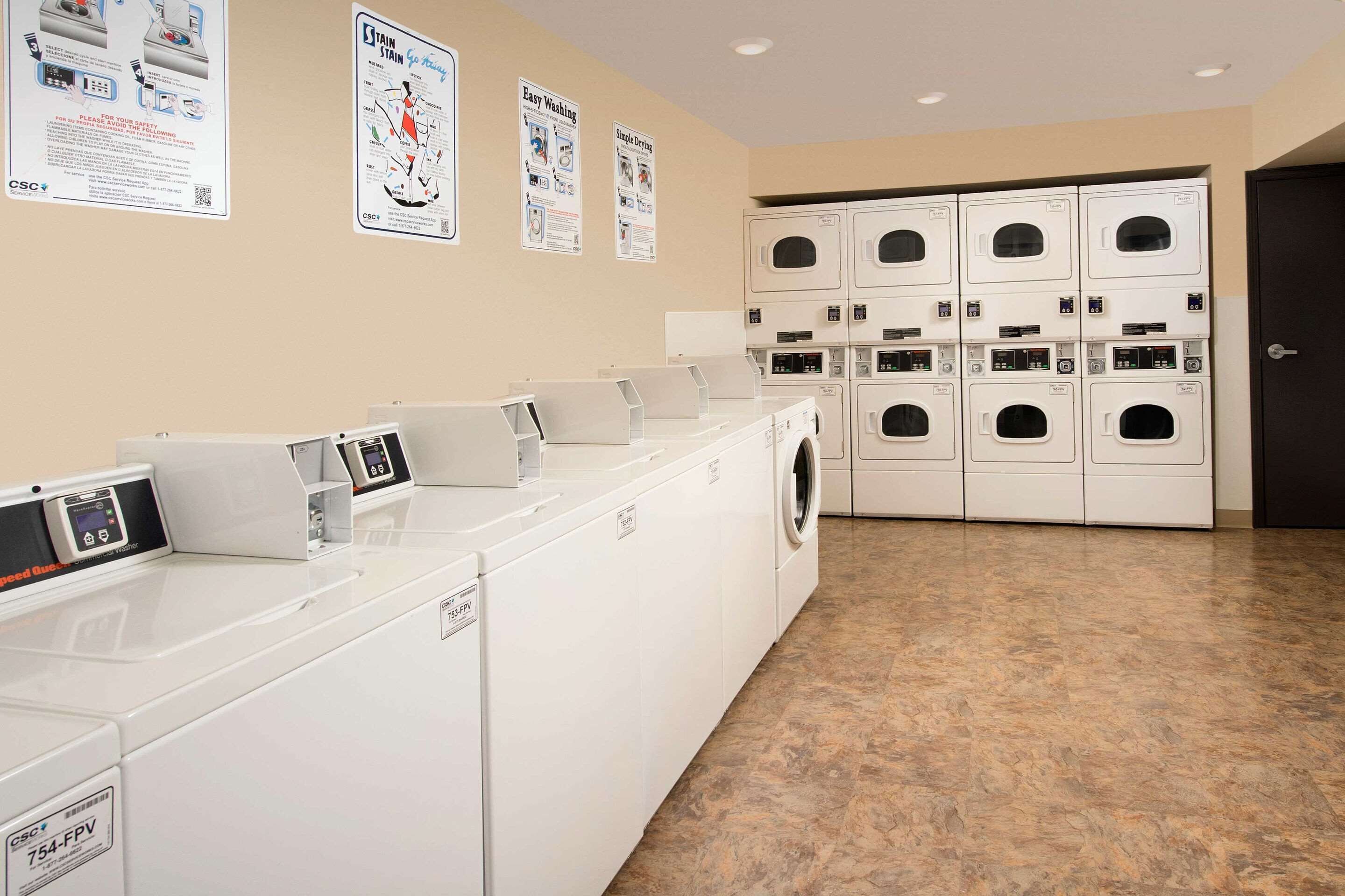 A laundry facility with multiple washing machines and dryers, a tiled floor, and instructional posters on the wall.