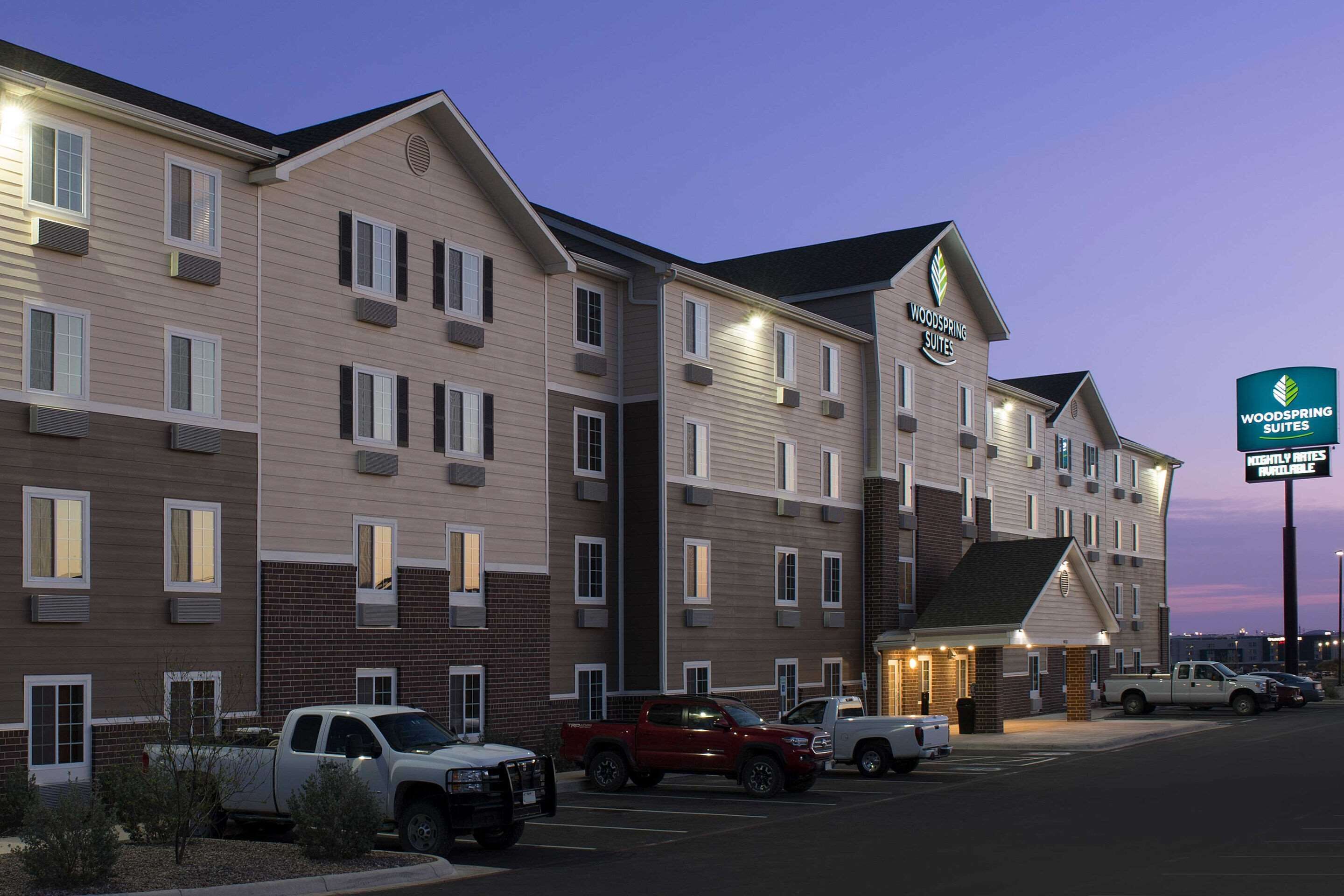 Um hotel de vários andares com telhado plano, uma placa indicando 'Woodspring Suites' e 'Tarifas Diárias Disponíveis', e vários veículos estacionados em frente.