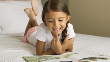 Un niño acostado en una cama con un libro, en una habitación con una colcha blanca y un cabecero de madera.