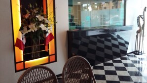 Uma área de recepção com piso xadrez, um balcão de atendimento e uma janela grande com vista para o exterior.