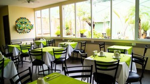Restaurante com várias mesas e cadeiras, grandes janelas com vista para o verde lá fora.