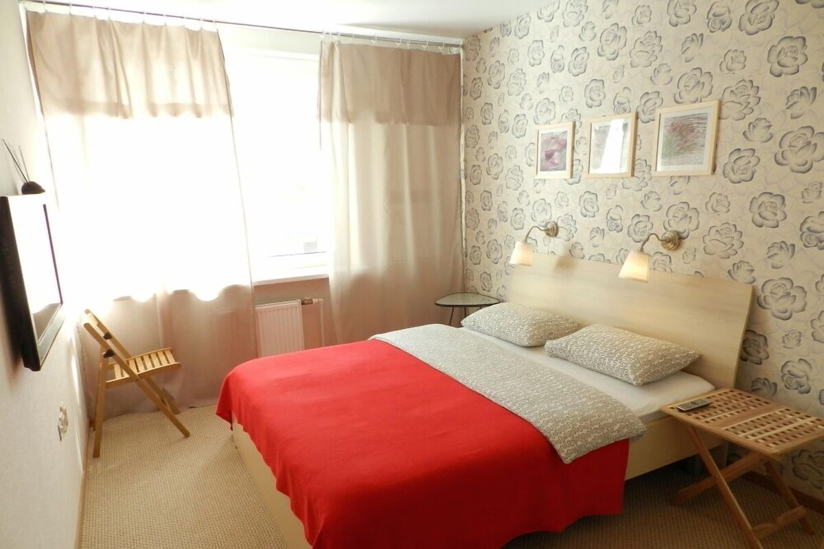 A bedroom with a bed, a nightstand, a lamp, a chair, and a window with curtains.