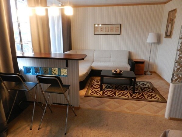 A compact living area with a sofa, a small table, and two chairs. A decorative rug is on the floor, and there is a framed picture on the wall.