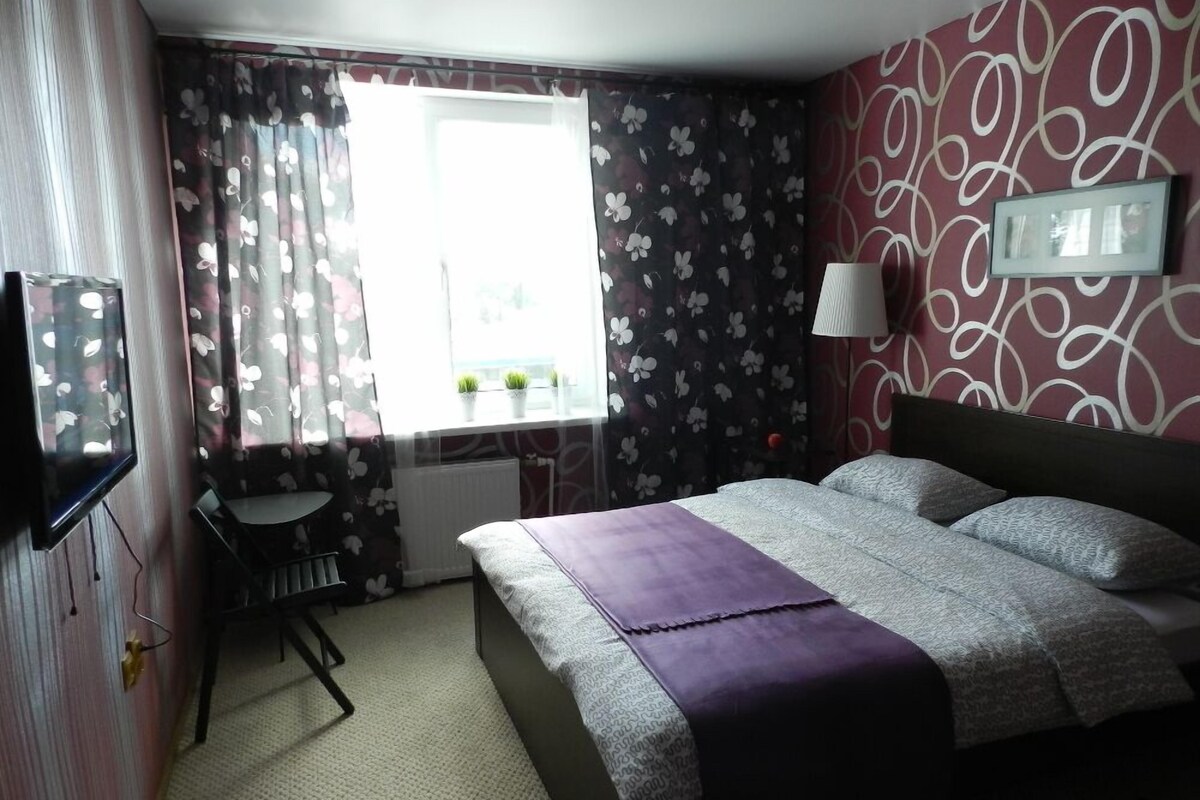 A hotel room with a bed, a desk, a chair, a window with curtains, a radiator, and a wall-mounted lamp.