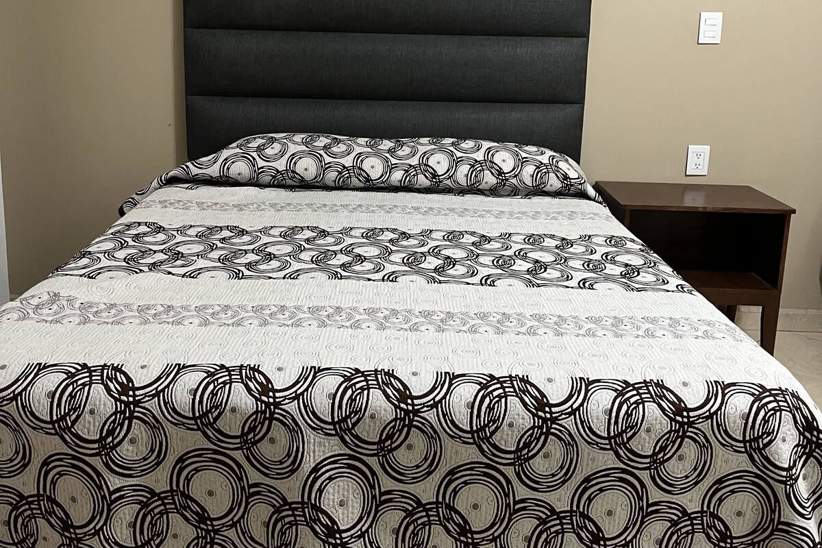 A bed with a patterned bedspread and pillows, a dark headboard, and a nightstand with a lamp.
