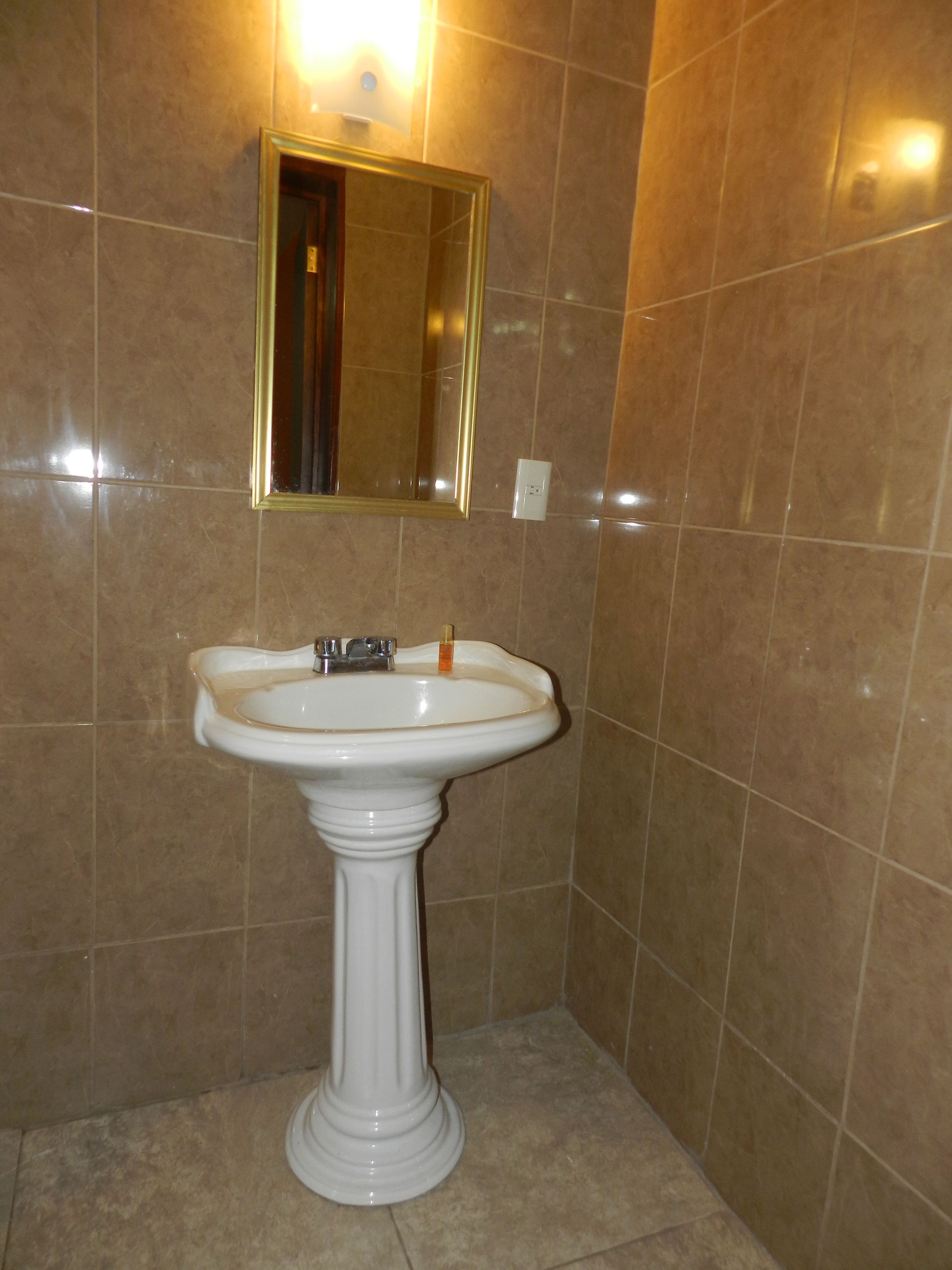 Een badkamer met een wastafel op een voetstuk, een spiegel en beige betegelde wanden.