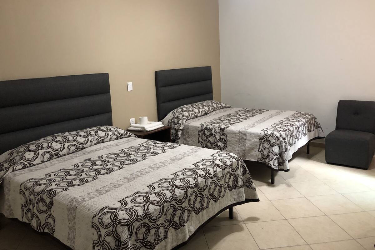 A hotel room with two beds, each with a headboard, a small table with a lamp, and a chair.