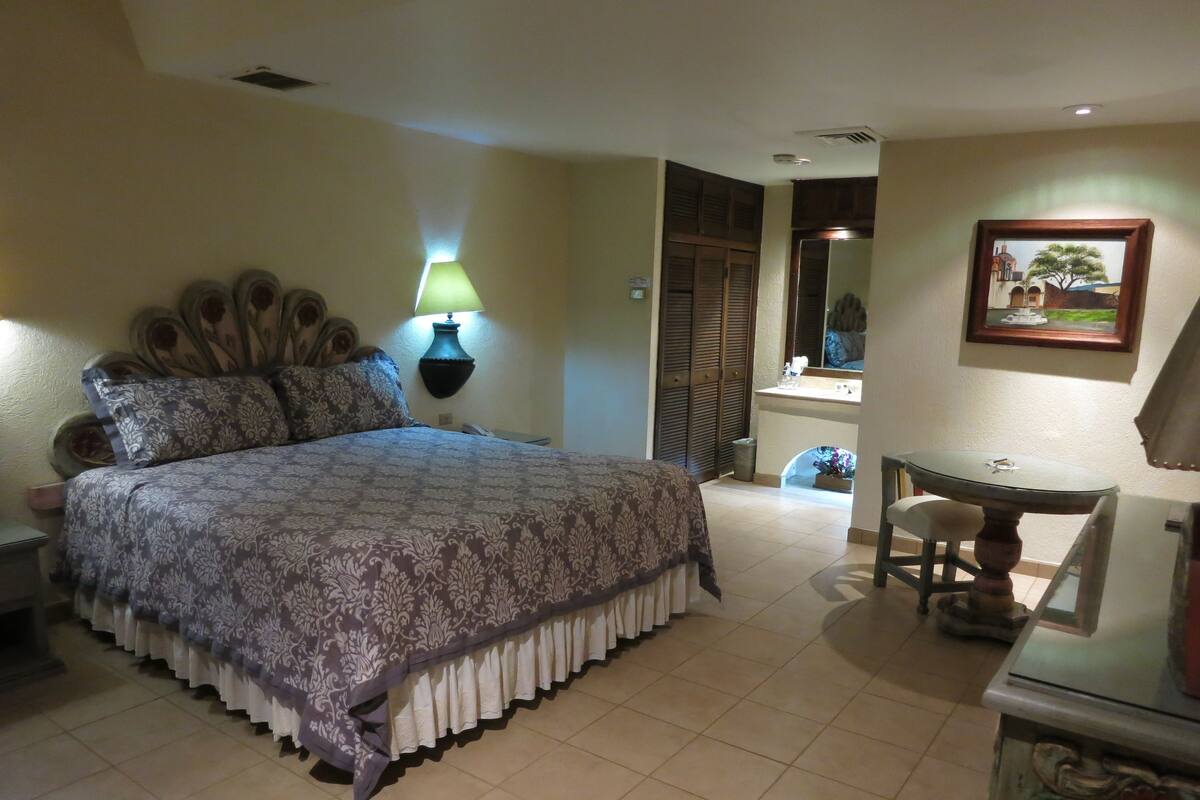 A bedroom with a large bed, a bedside table, a lamp, a mirror, a small table, and a chair.