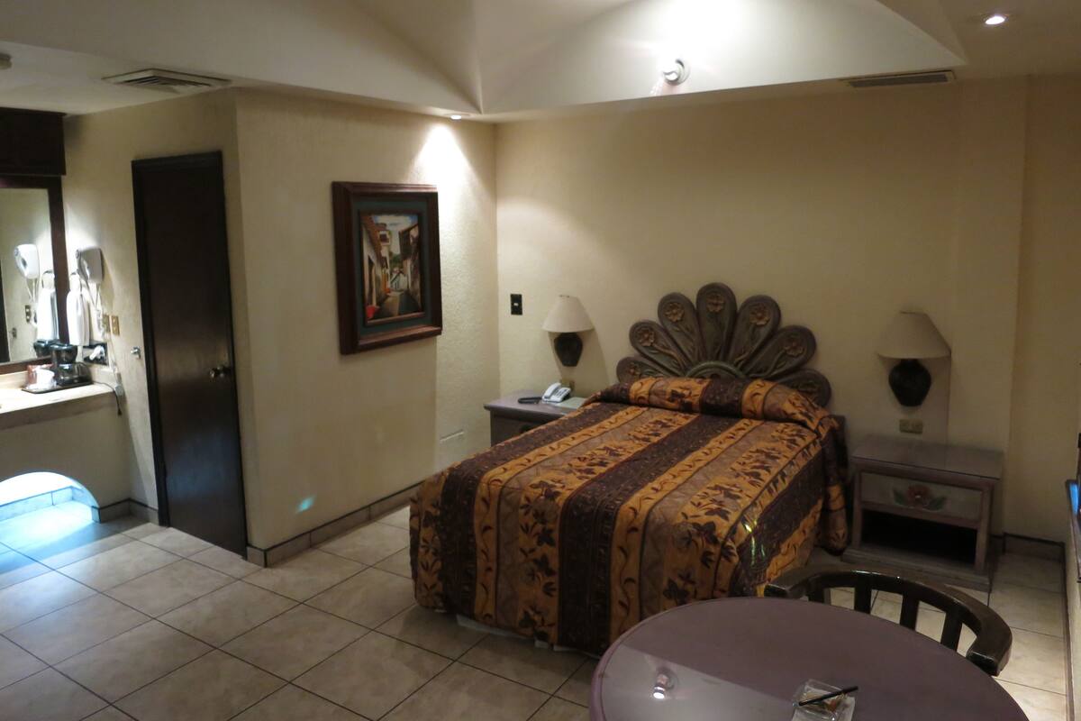 A hotel room with a bed, bedside lamps, a desk, a chair, a mirror, and a painting on the wall.