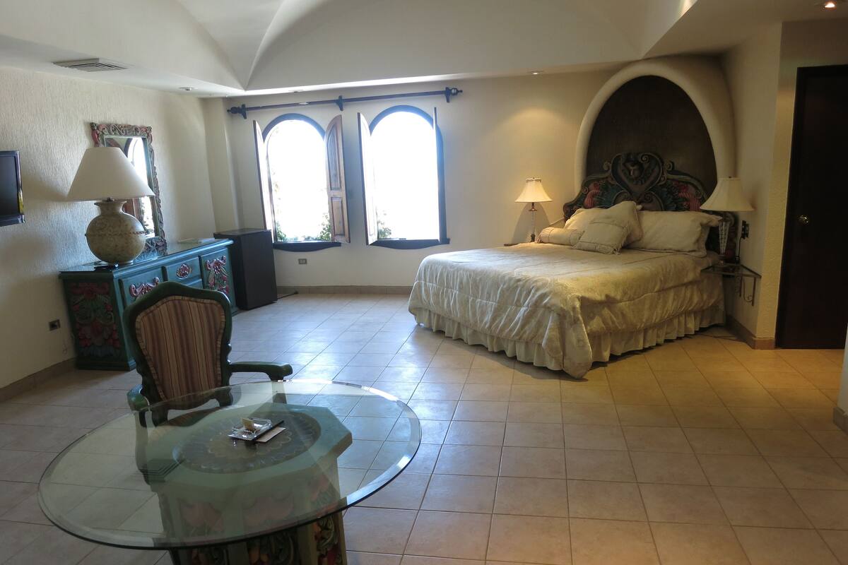 A bedroom with a bed, a glass table, a chair, a dresser, and two windows with curtains.