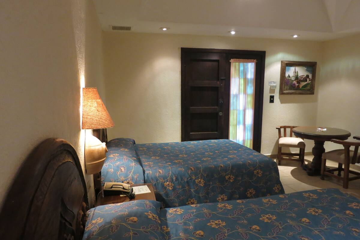 A hotel room with two beds, a desk, a chair, a lamp, and a door.