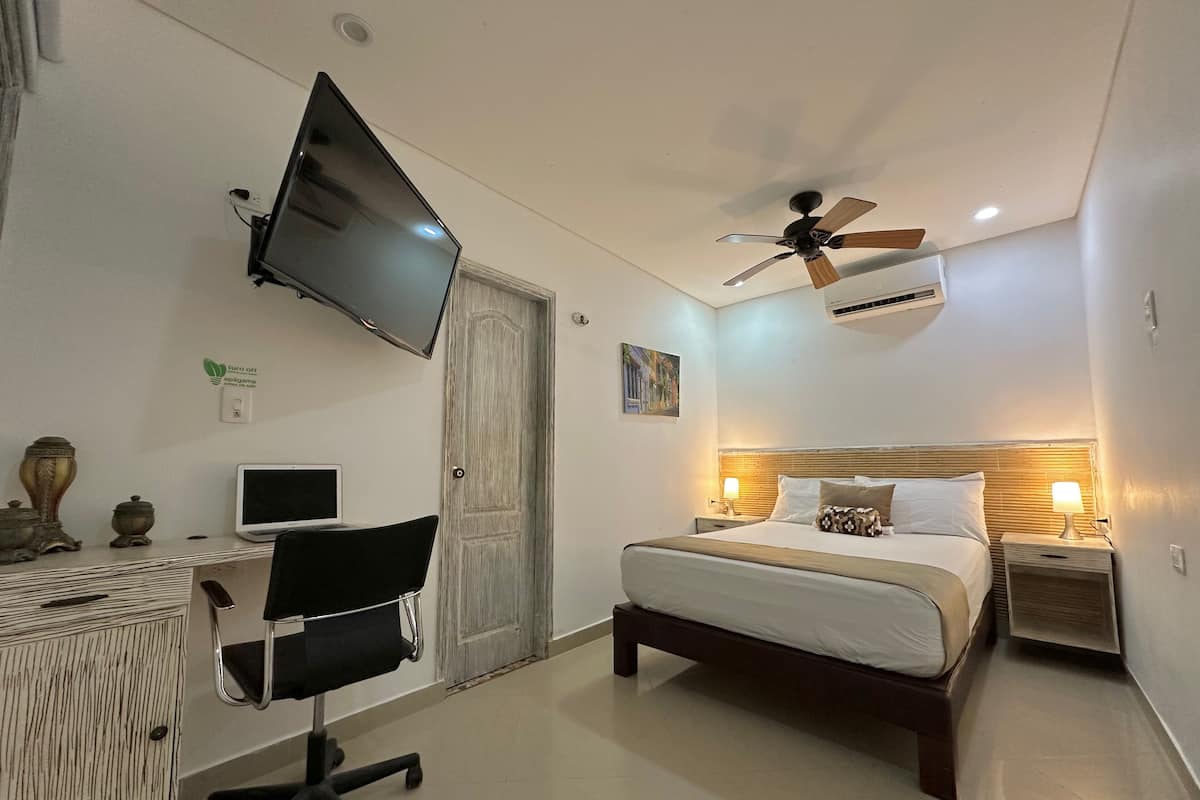 A bedroom with a bed, bedside lamps, a desk with a chair, a TV, a ceiling fan, and an air conditioning unit.