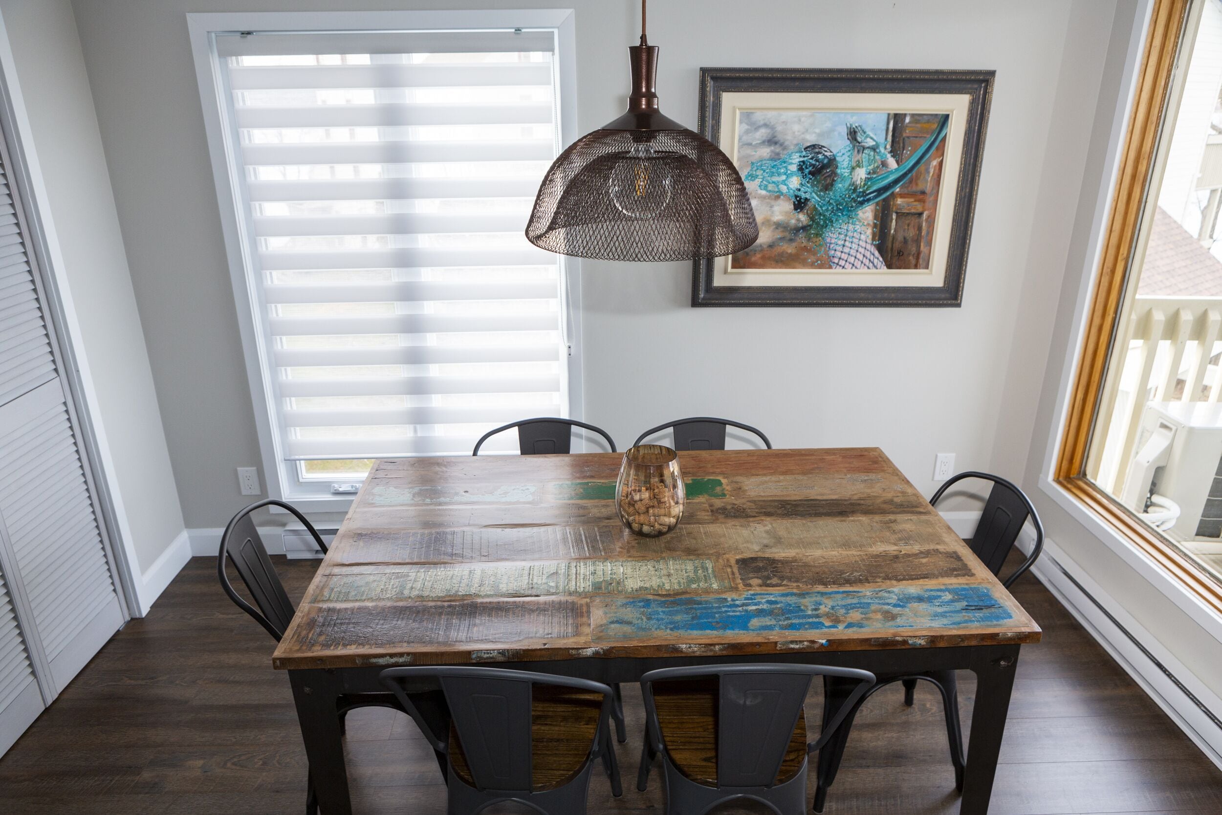 A dining area with a rustic wooden table, black chairs, a hanging pendant light, and a framed painting on the wall.