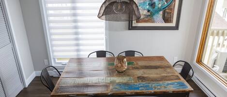 A dining area with a rustic wooden table, black chairs, a hanging pendant light, and a framed painting on the wall.