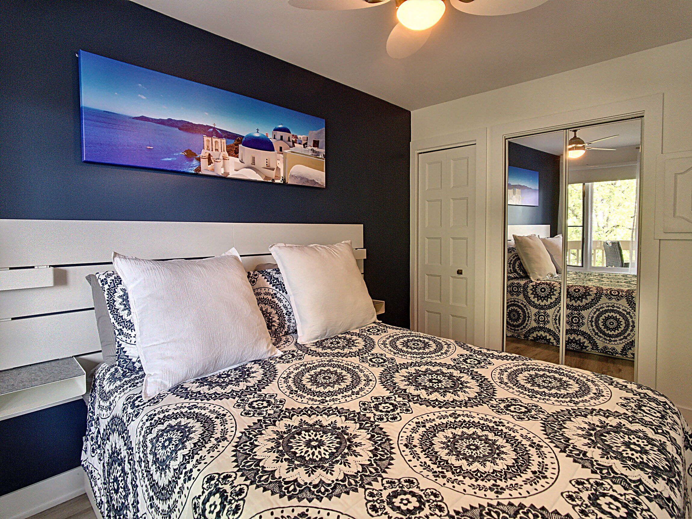 A bedroom with a large bed, a headboard, a painting of a coastal scene, and a door leading to another room.