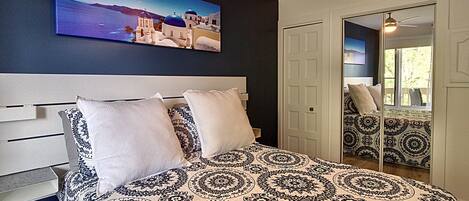 A bedroom with a large bed, a headboard, a painting of a coastal scene, and a door leading to another room.