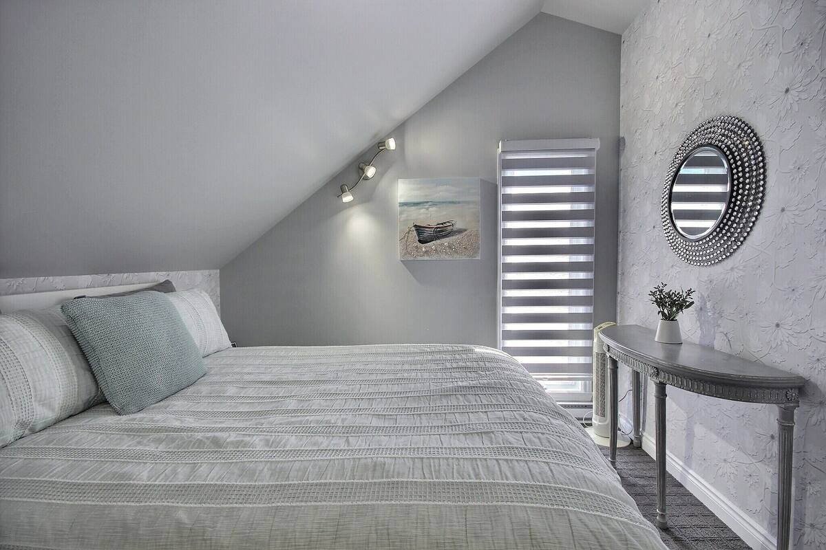 Une chambre avec un plafond en pente, un lit recouvert d’une couvre-lit à rayures, une table de chevet avec un miroir et un tableau accroché au mur.