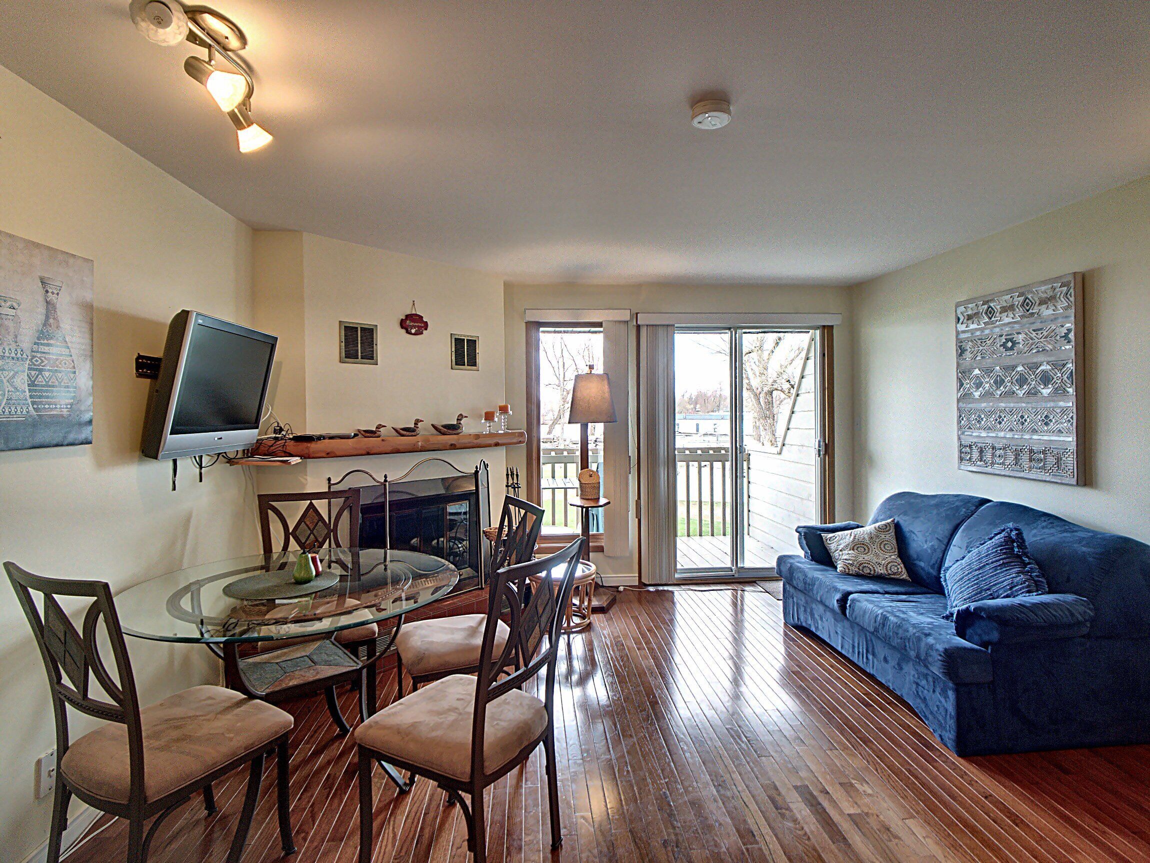 A living room with a glass dining table, wooden chairs, a blue sofa, and a television on the wall.