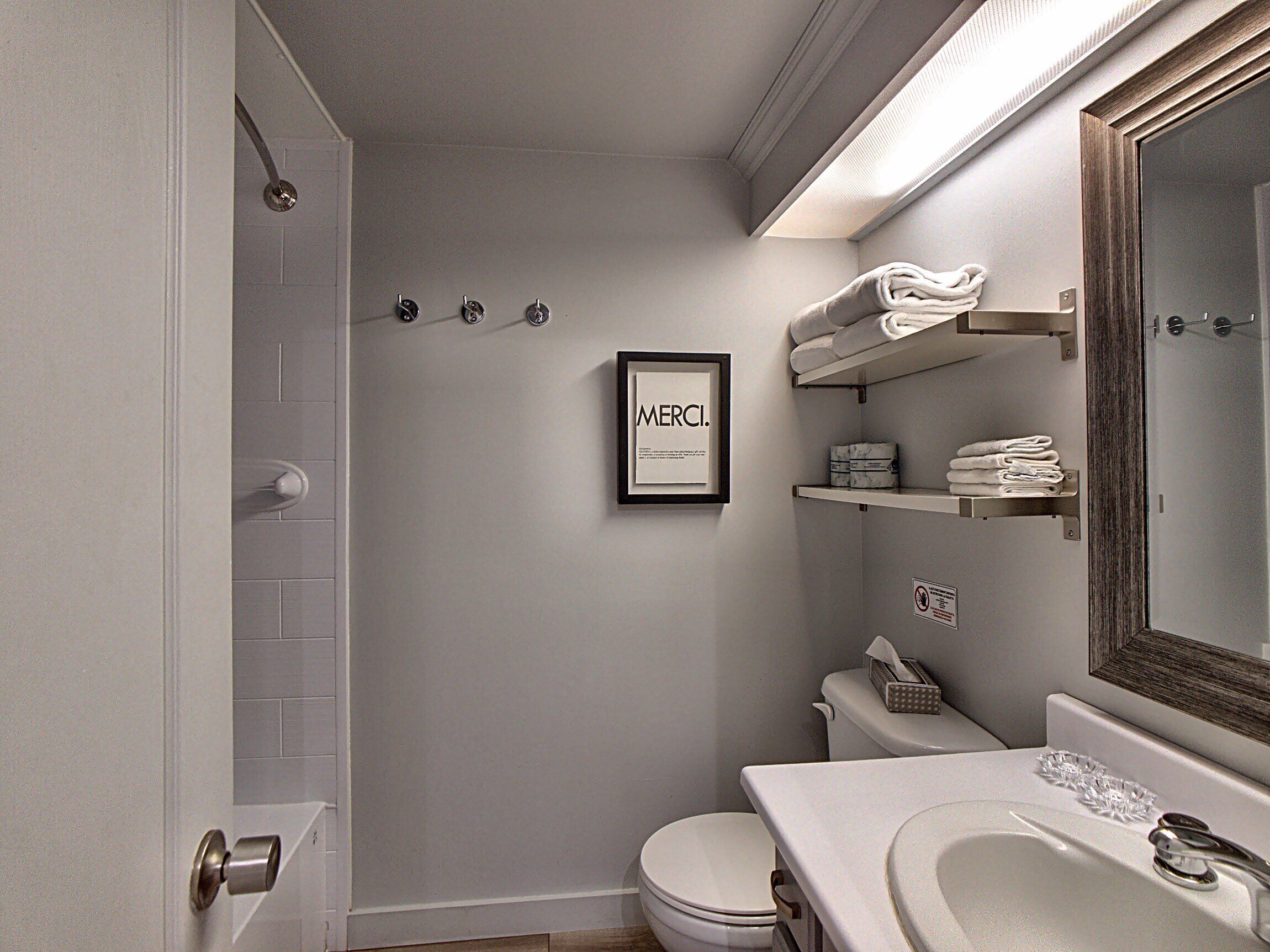 A modern bathroom with a toilet, sink, and shower. There is a framed sign on the wall and towels on a shelf.