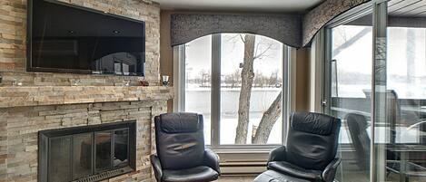 Una sala de estar moderna con chimenea de piedra, sillones de cuero, televisor de pantalla plana y un amplio ventanal con vista a un lago.