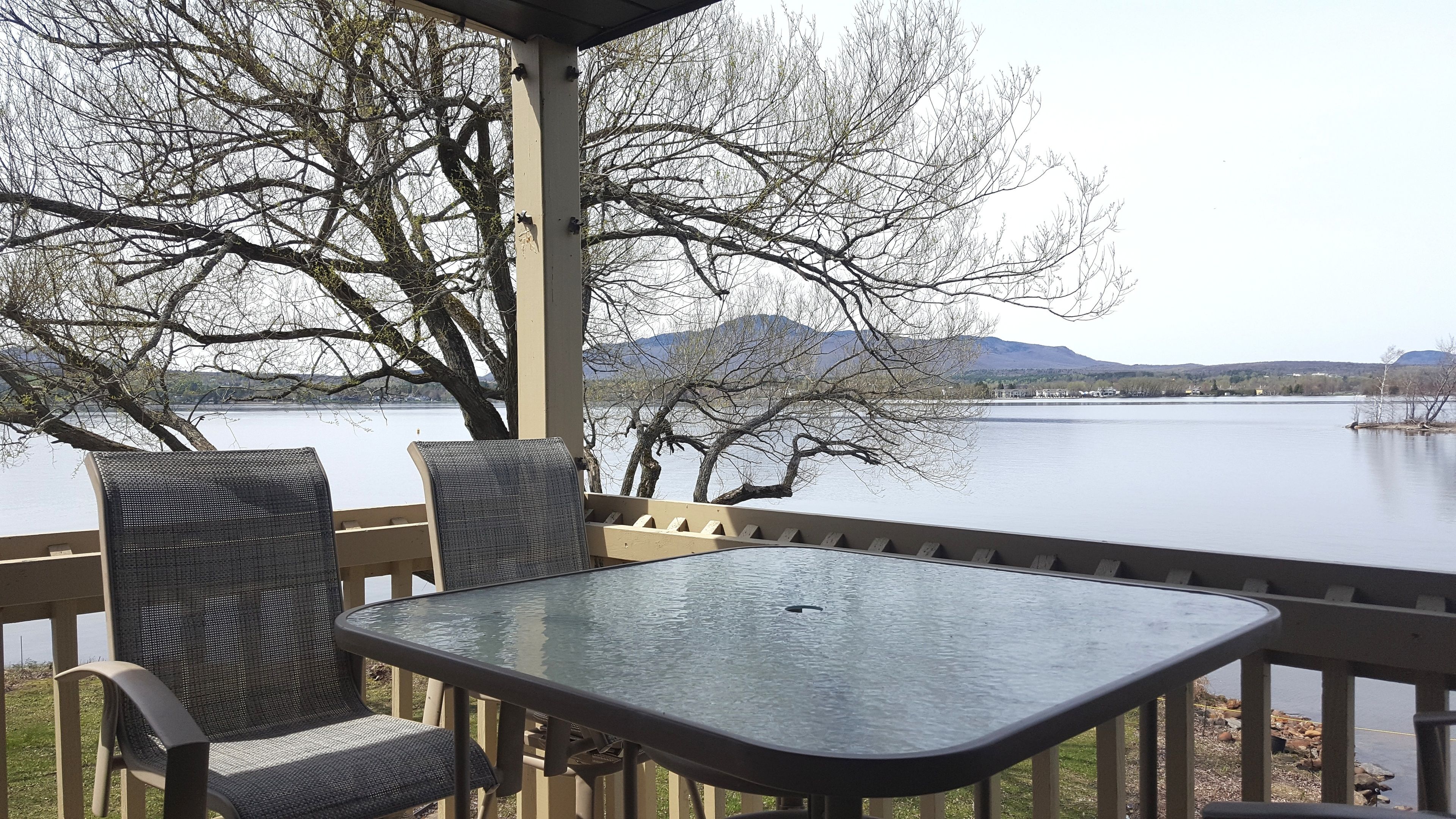 A covered outdoor seating area with a table and chairs, overlooking a calm lake with trees and distant mountains.