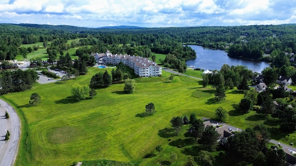 An aerial view of a large, multi-story hotel with a swimming pool, surrounded by lush green fields and a lake. - Condo 2 Bedrooms Jardin des Sables (Orford)
