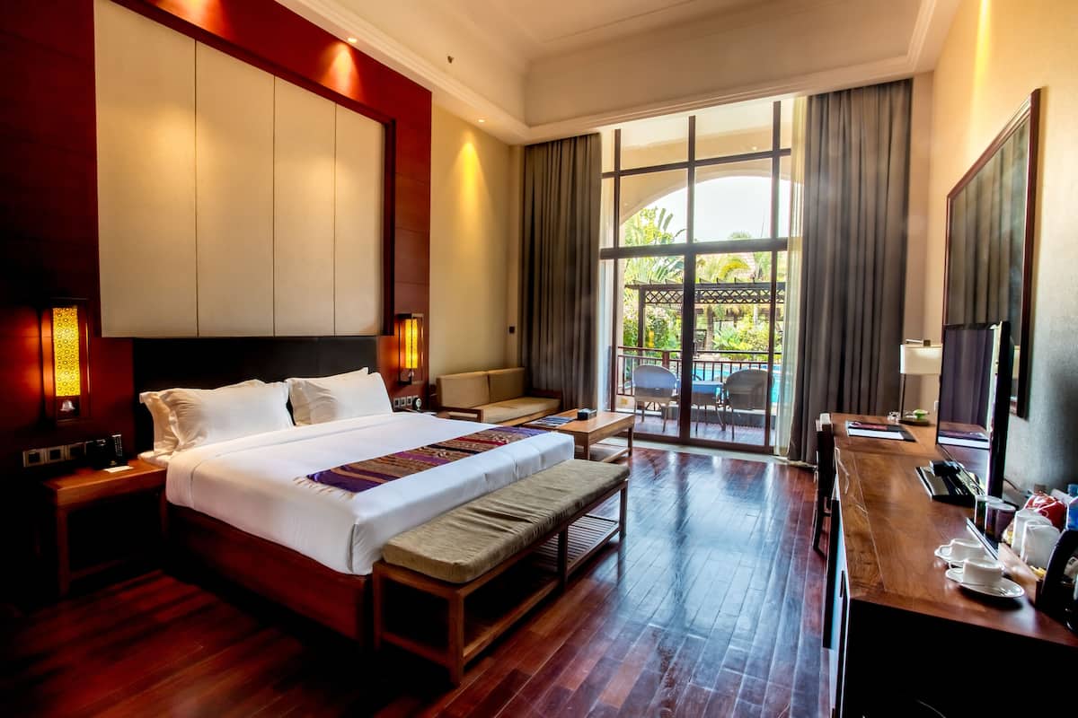 Ein Hotelzimmer mit einem großen Bett, einer Holzbank, einem Schreibtisch mit Stuhl und Blick ins Freie durch große Fenster.