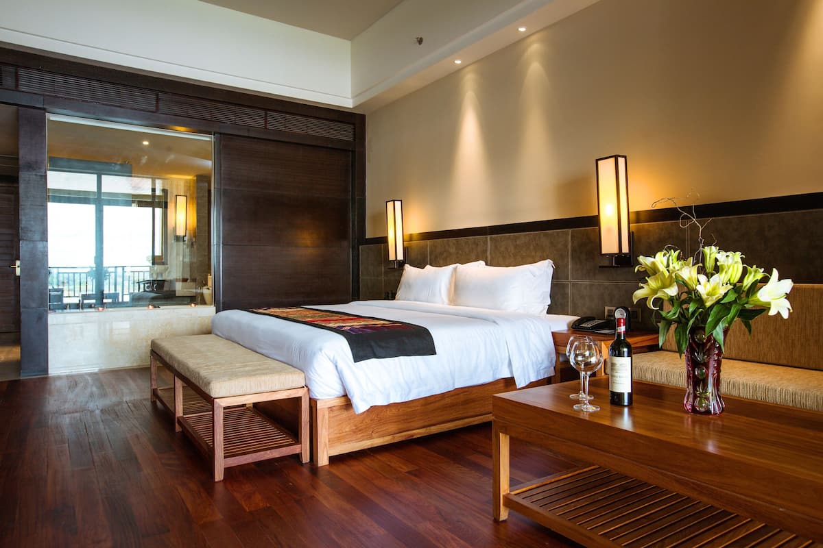 Ein Hotelzimmer mit einem großen Bett, einer Holzbank, einem Tisch mit Blumenvase und Blick auf den Balkon.