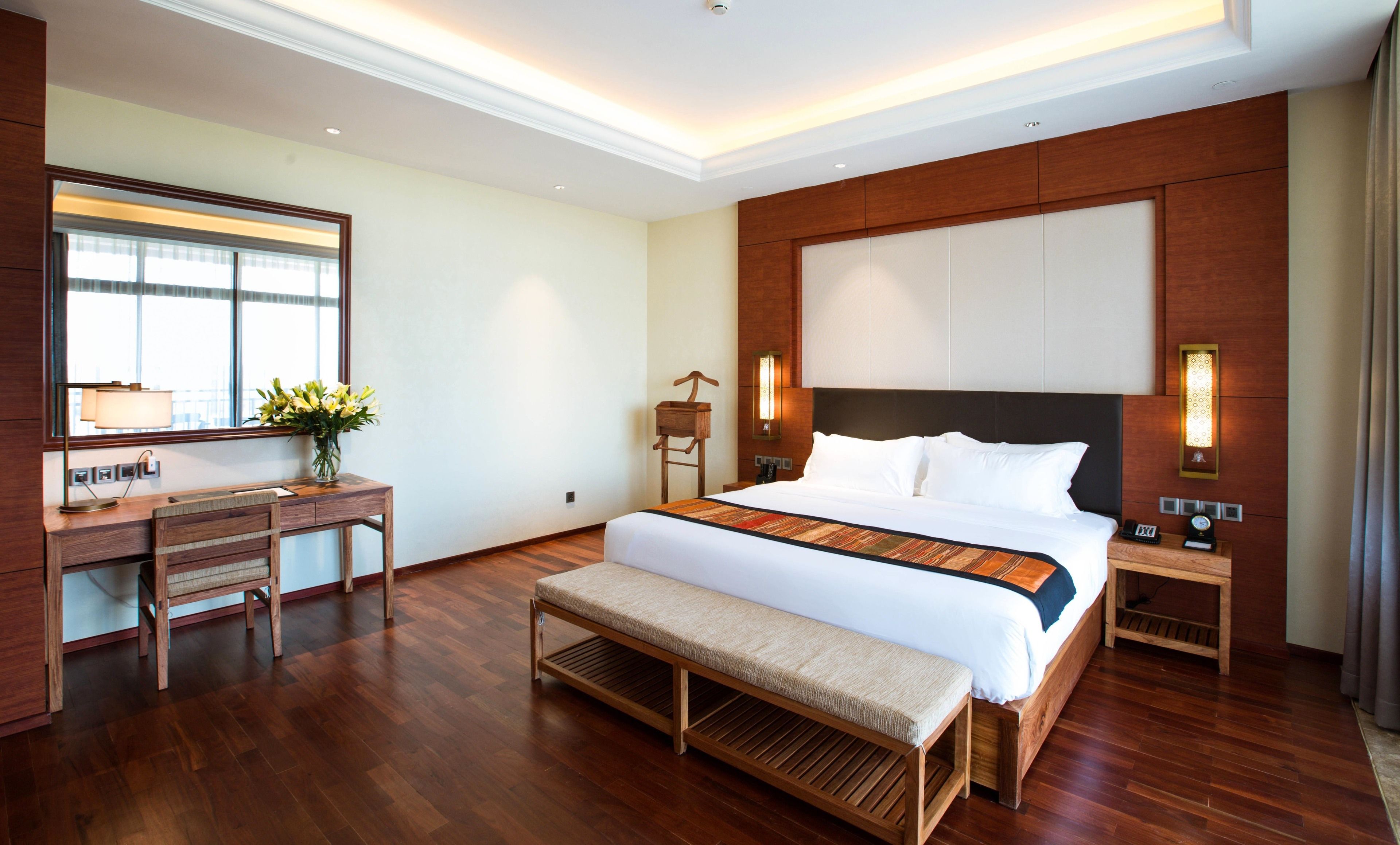 A hotel room with a large bed, a desk with a lamp, a bench, and a mirror.