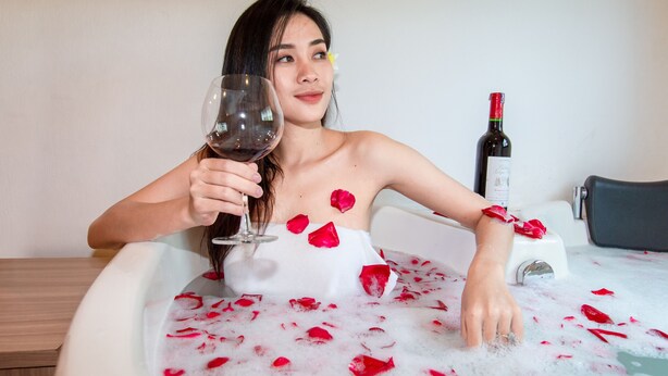 Une personne dans une baignoire remplie d’eau et de pétales de rose, tenant un verre de vin.