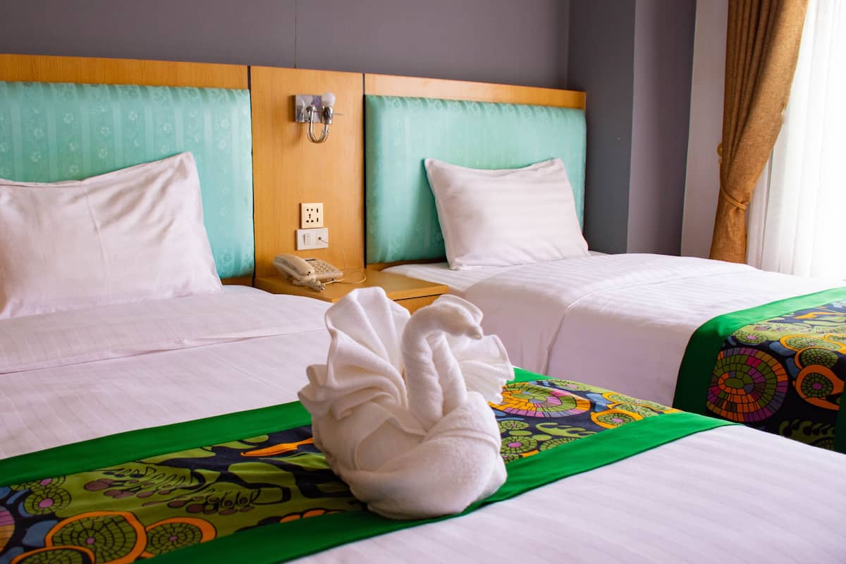 Quarto de hotel com duas camas, cada uma com roupa de cama branca e colcha verde estampada. Tem uma cabeceira de madeira com um telefone e uma tomada.