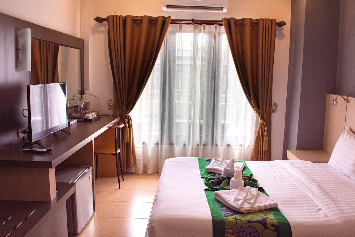 Quarto de hotel com cama, escrivaninha com computador, televisão e janela com cortinas.
