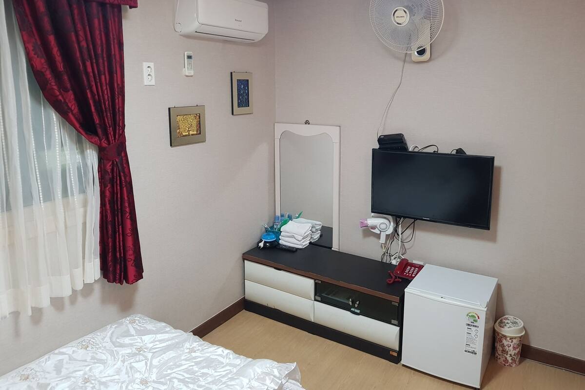 Et hotelværelse med seng, sminkebord med spejl, et TV ophængt på væggen, et lille køleskab og en skraldespand.