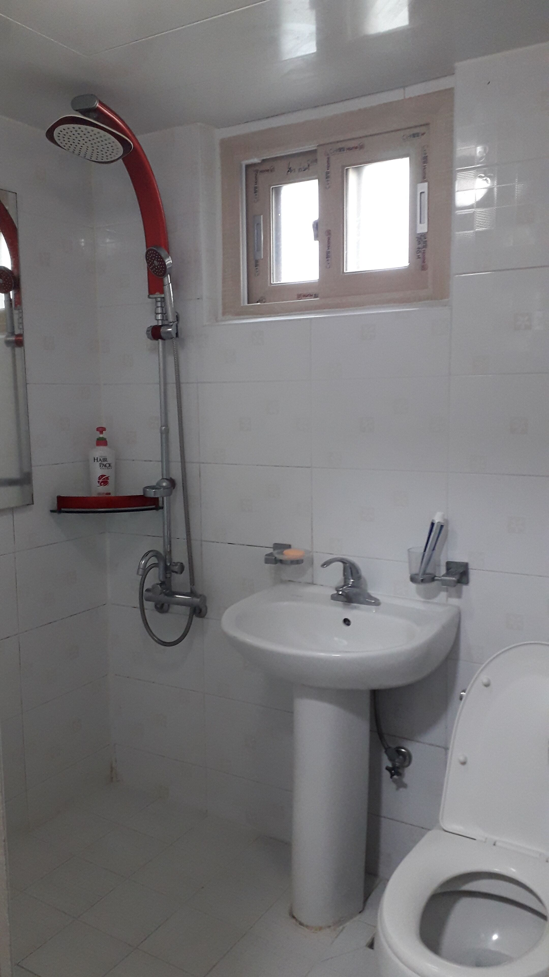 A bathroom with a white sink, a toilet, a shower with a red handle, and a window above the sink.
