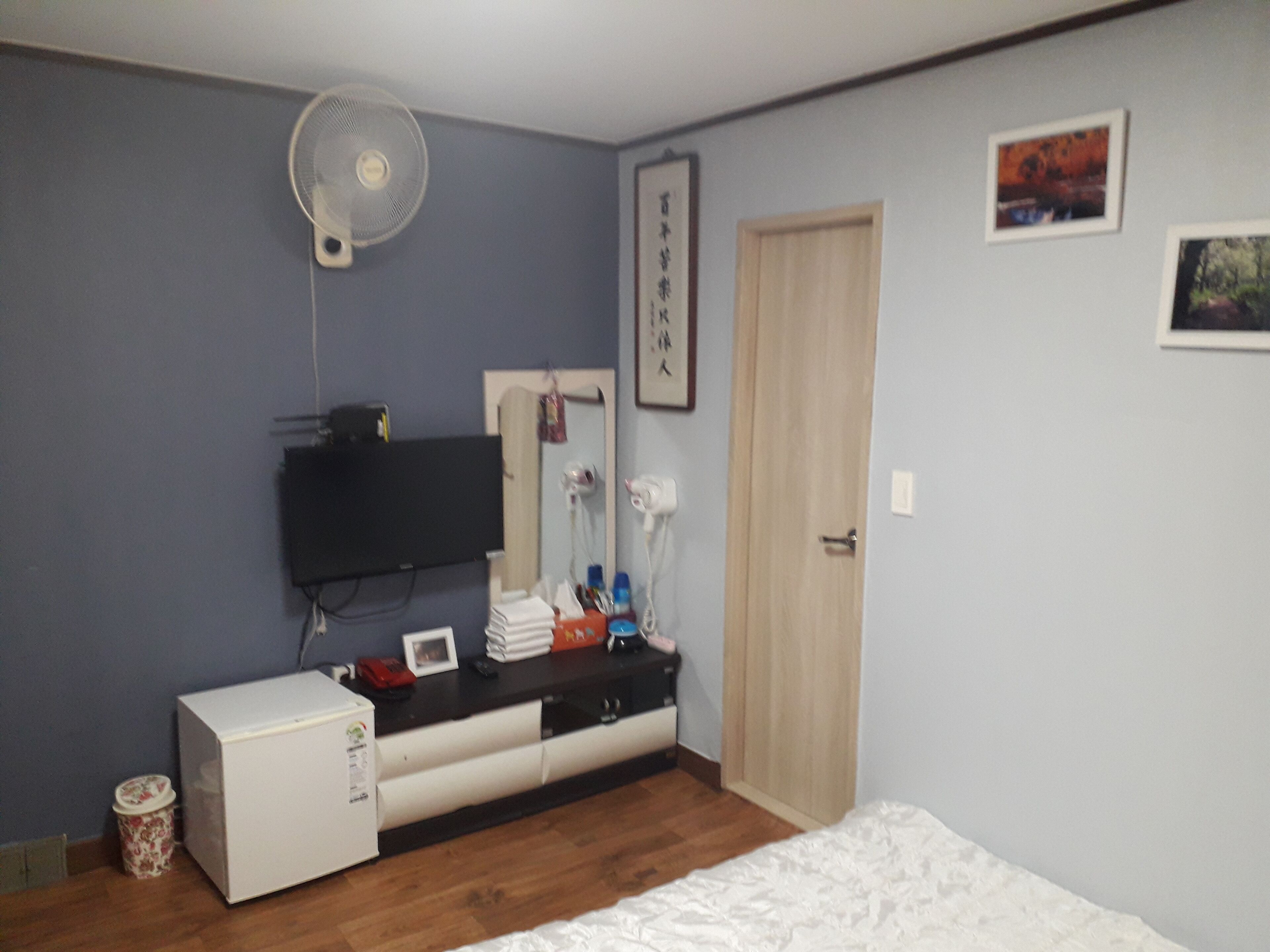 A bedroom with a bed, a small white refrigerator, a desk with a computer, a wall-mounted fan, a door, and a framed picture.