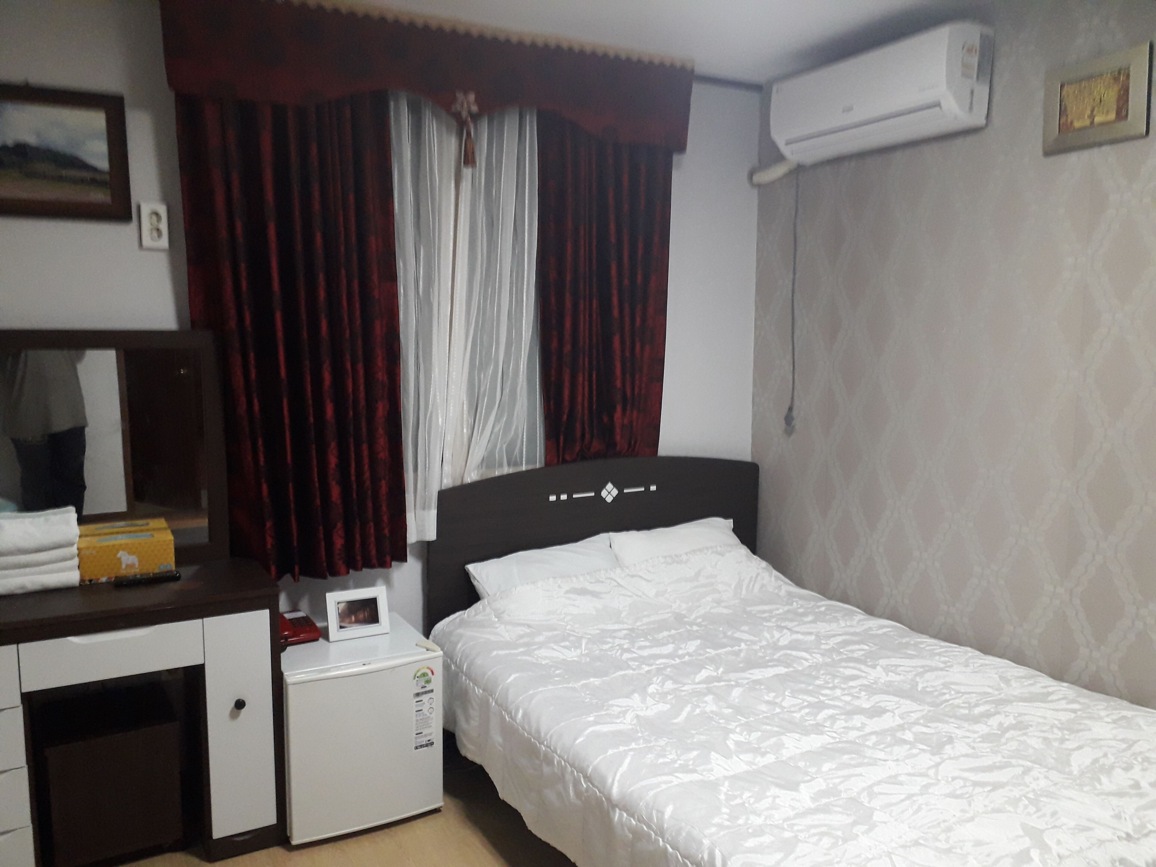 A hotel room with a bed, a desk with a microwave, a small refrigerator, a mirror, and a window with curtains.