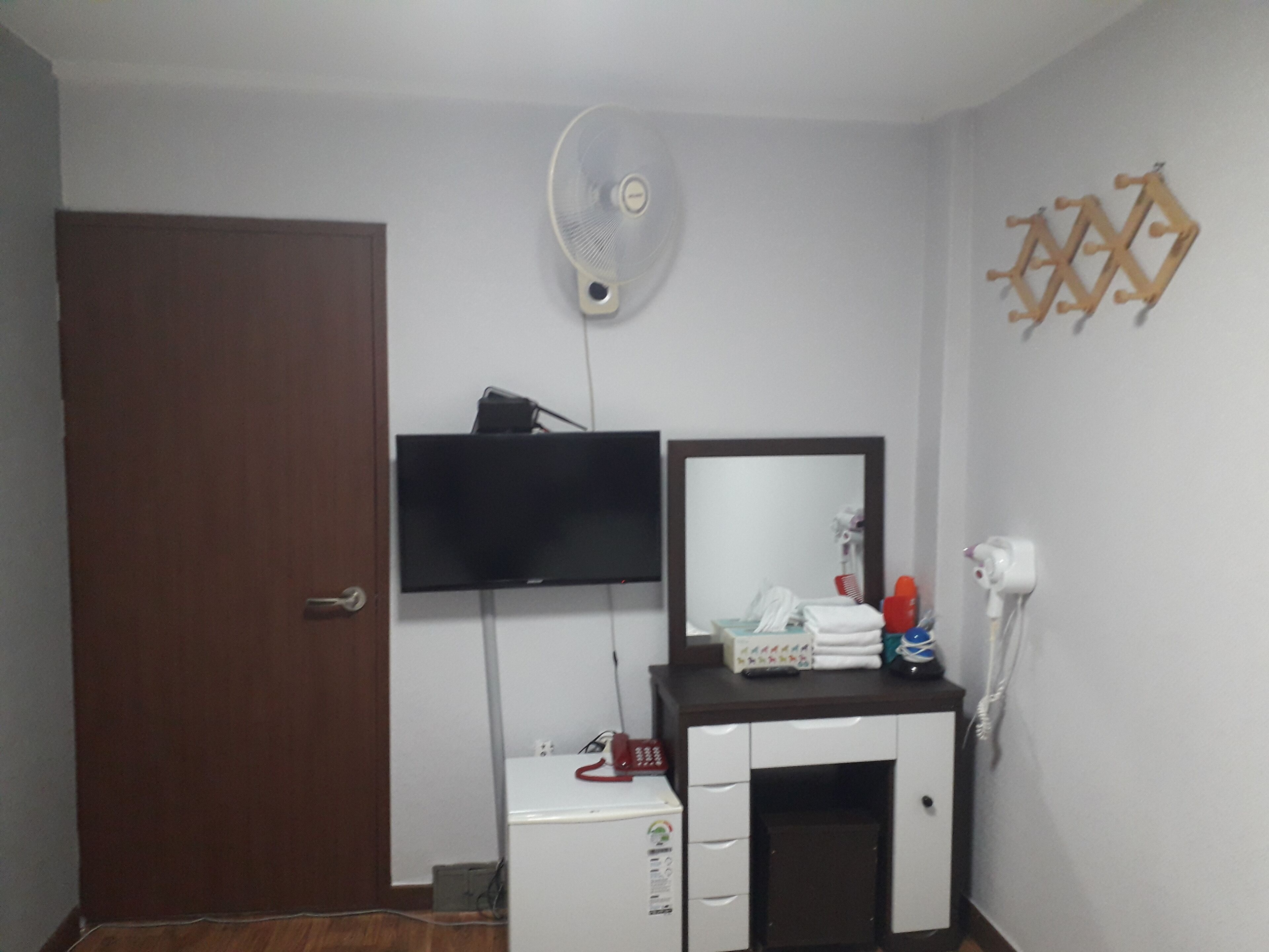 Een kamer met een houten deur, een witte kast met spiegel, een televisie en een ventilator aan de muur.