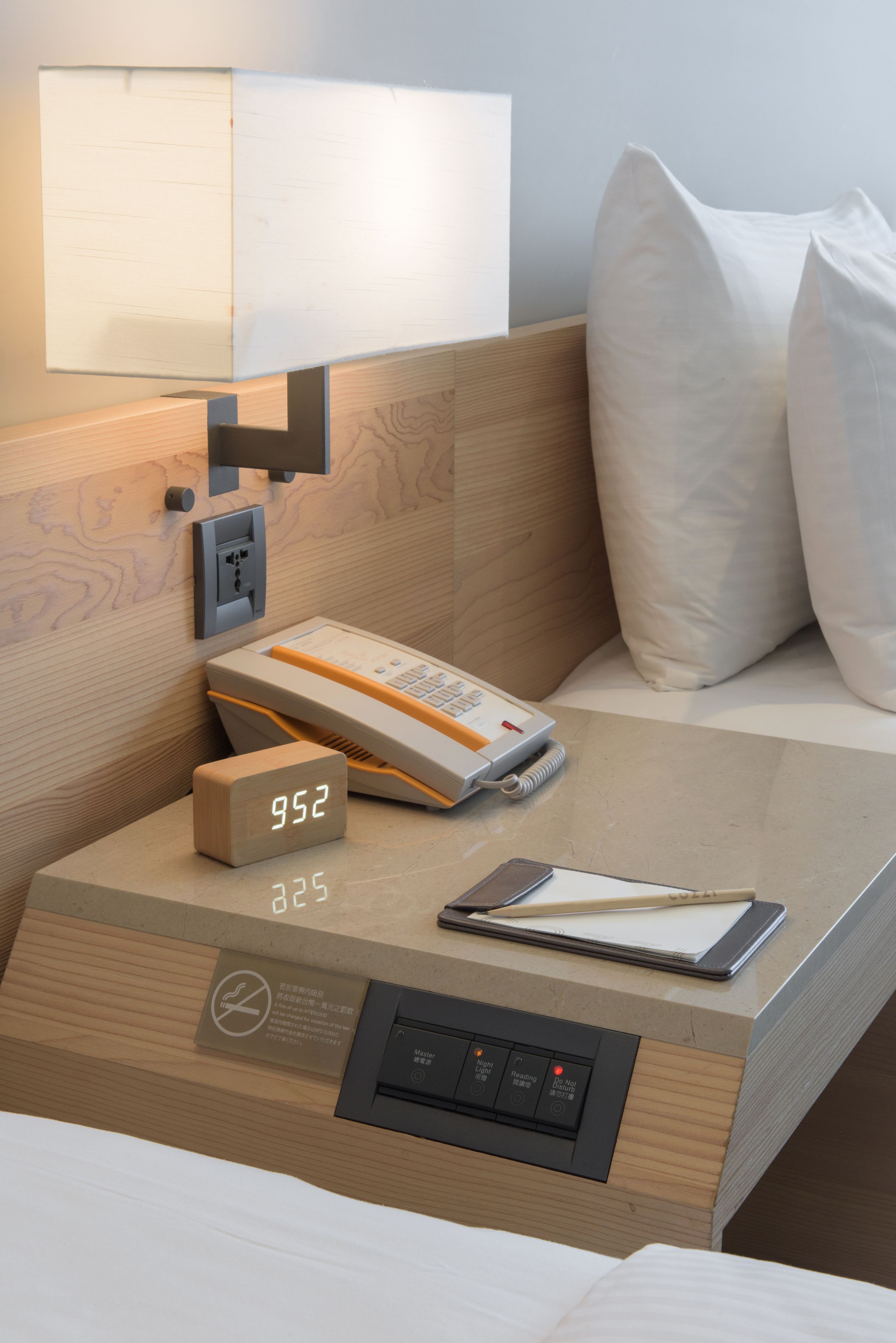 A neatly arranged hotel room with a wooden headboard, a bedside table with a digital clock, a telephone, and a notepad.